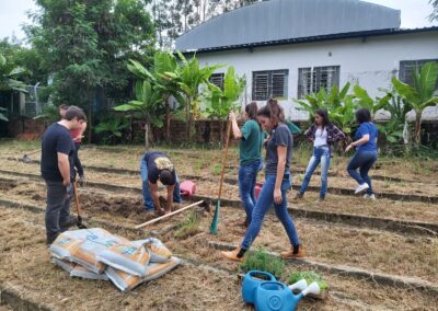 Seis alunos trabalhando na horta, espalhados pelo local. Um está agachado, outra está preparando o solo. E o resto está dividido, auxiliando.